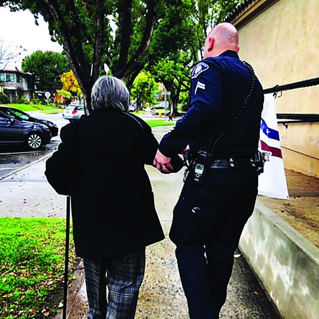 Officer assists elderly person with cane on sidewalk near building; trees, parked cars, and overcast sky visible in background