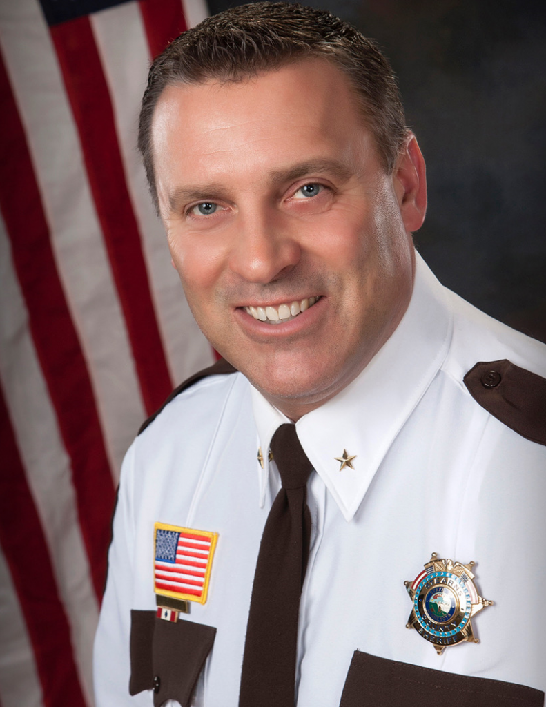 Uniformed sheriff’s official wears a white dress shirt with brown shoulder panels, tie, gold star badge, collar stars, U.S. flag patch, and service ribbon, posed before a U.S. flag and studio backdrop