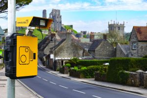 Yellow speed enforcement camera mounted on a pole overlooks a two‑lane road, with village houses, stone buildings, rooftops, and a ruined castle on a hill