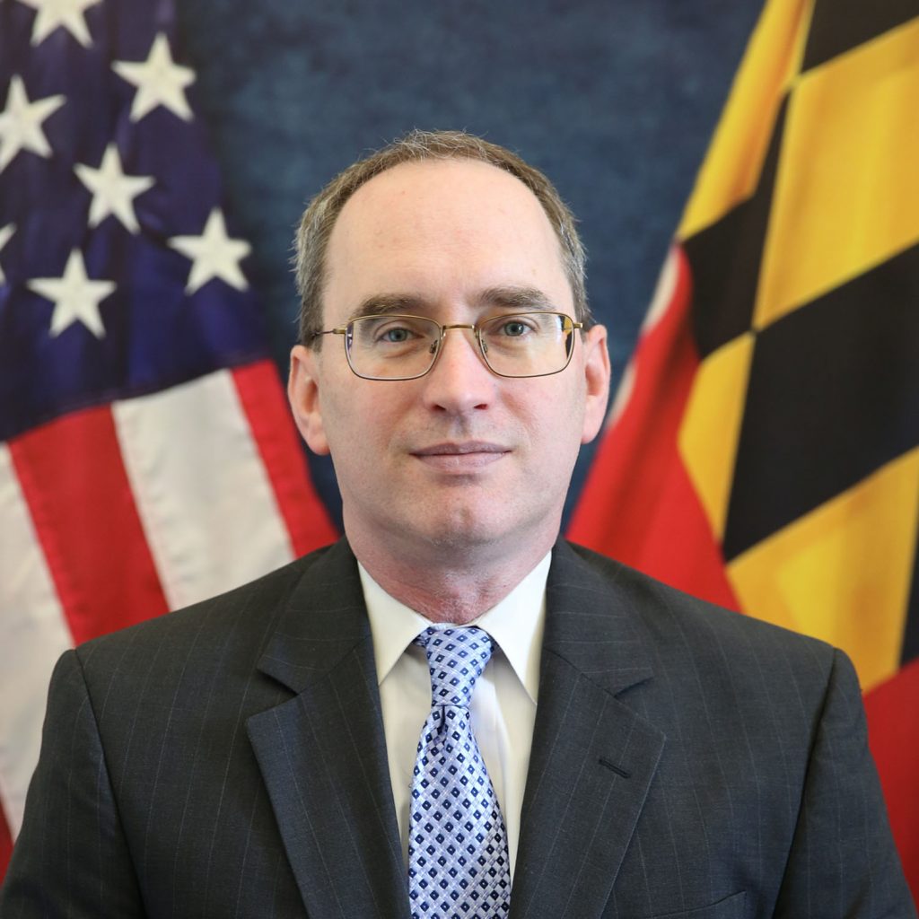Man in dark pinstripe suit and blue patterned tie stands before U.S. and Maryland flags; both flags fully visible and prominently displayed behind him