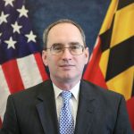 Man in dark pinstripe suit and blue patterned tie stands before U.S. and Maryland flags; both flags fully visible and prominently displayed behind him
