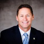 a person wearing a dark pinstripe suit, a white dress shirt, and a light blue tie with small white patterns. The person has short brown hair and is wearing an enamel lapel pin on the left side of the suit jacket. The background is a plain gray gradient.