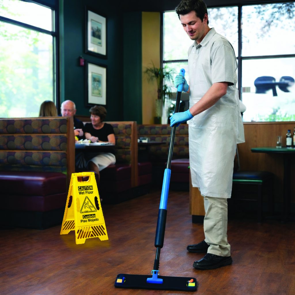Custodial worker uses a blue spray mop on a restaurant floor, wearing gloves and apron, with a yellow ‘Caution Wet Floor’ sign, booths, tables, and seated patrons in the background