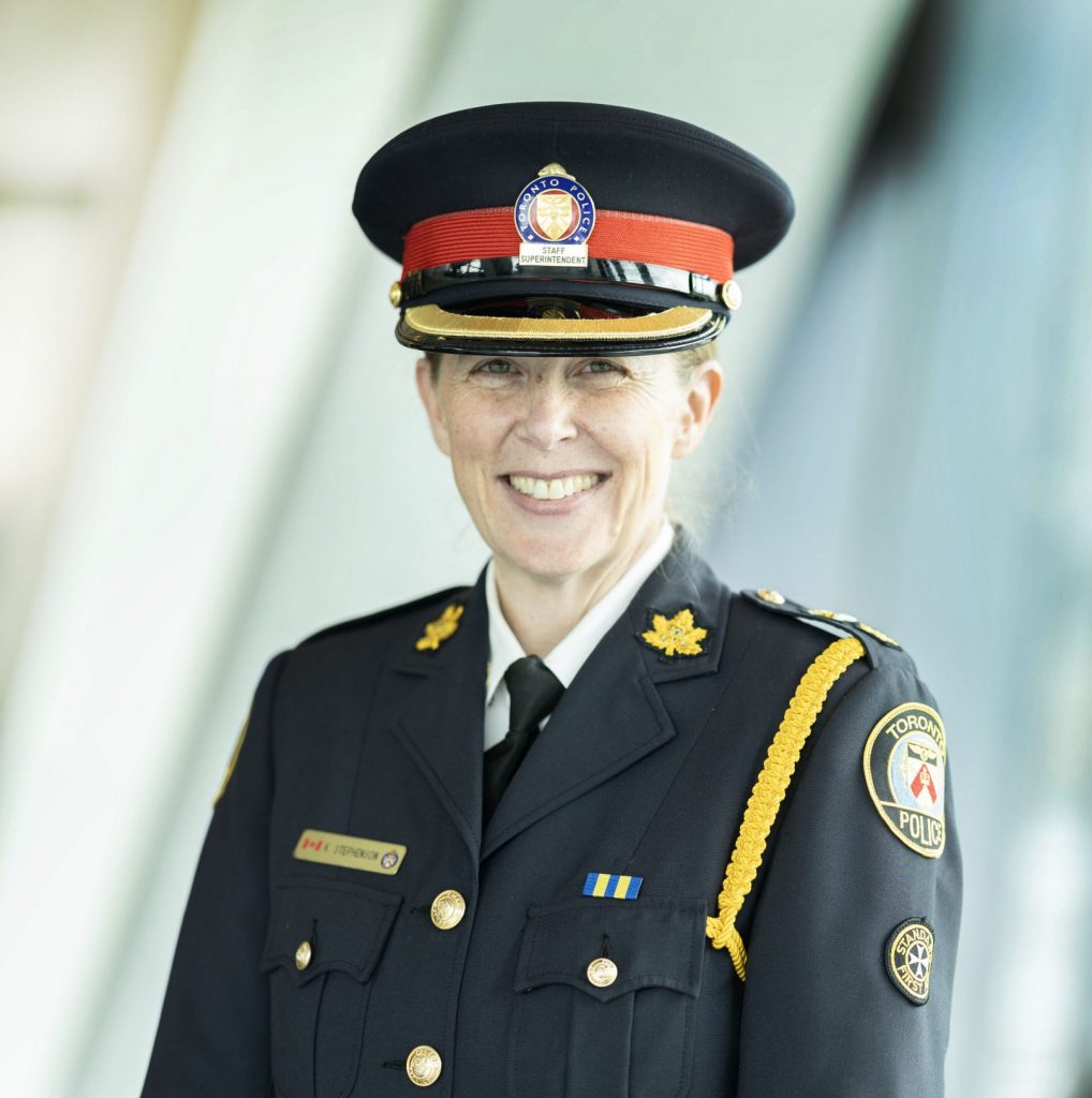 Formal portrait of a senior police officer wearing a dark dress uniform with peaked cap, gold cords, badges, ribbon bar, and shoulder patches, standing before a modern glass‑walled interior