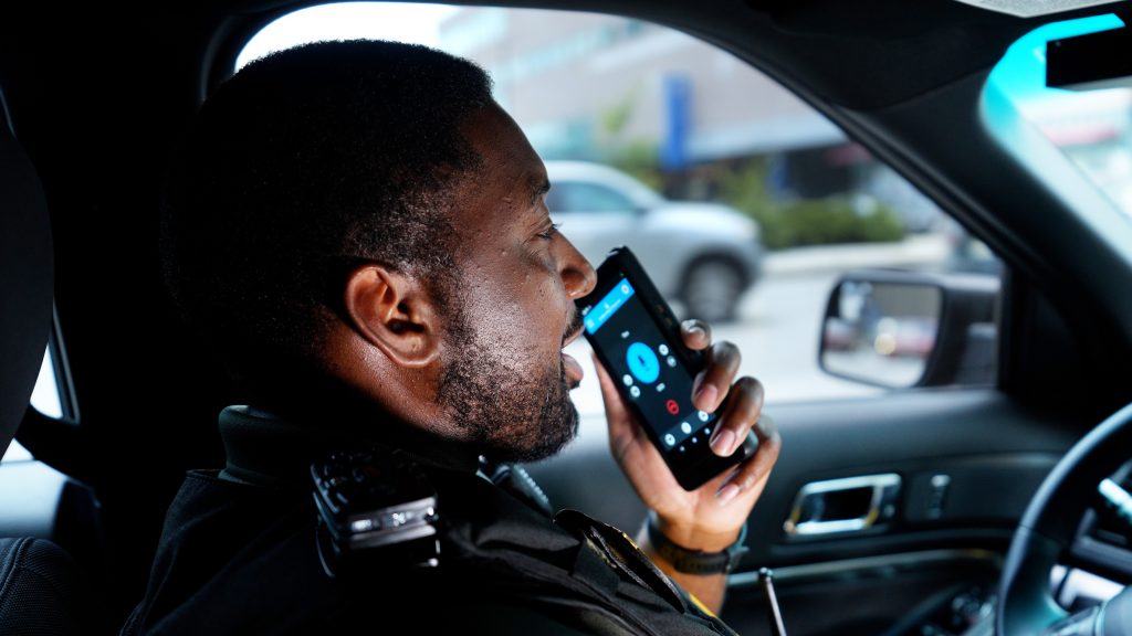 A law enforcement officer is seated in the driver's seat of a patrol vehicle. The officer is holding a smartphone to the ear with the screen visible, showing an active call interface. The interior of the vehicle includes standard equipment such as a radio microphone attached to the shoulder and part of the dashboard and steering wheel are visible. Outside the vehicle, there is a blurred view of buildings and greenery, indicating an urban environment.