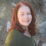 a person with shoulder-length, wavy red hair standing outdoors. The background consists of dry grass and rocks. The person is wearing a green knit sweater and a silver necklace with a pendant.