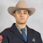 officer from the chest up, wearing a formal dark blue uniform with red piping on the shoulders. The uniform includes a light brown shirt and a blue tie. On the left side of the chest, there is a name tag that reads "SANDERSON." On the right side of the chest, there is a circular badge with an emblem in its center. The officer is wearing a beige cowboy hat. The shoulder patch on the left arm features intricate designs and text that are not fully legible in this image. The officer's face is visible, showing a neutral expression. The background is plain and light gray