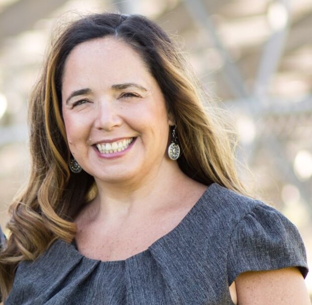 Person with long wavy hair wearing a gray pleated dress and dangling earrings stands outdoors against a blurred metal structure in bright natural light.
