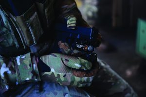 Close-up of a person in camouflage uniform and gloves holding a black handgun equipped with a mounted tactical light and laser attachment