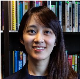 Individual wearing a dark blazer over a dark top sits before bookshelves filled with books and binders, with library shelving, colorful spines, and reference materials visible throughout the background
