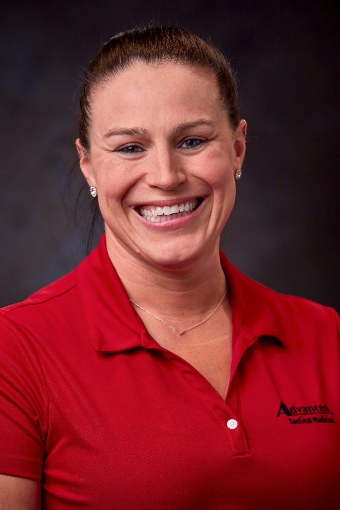Studio portrait of an individual wearing a red short‑sleeve polo shirt with an embroidered company logo, a necklace, and small earrings, posed against a dark neutral background under soft lighting