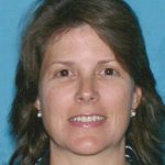 Individual with shoulder-length brown hair in black and white striped shirt stands before blue backdrop.