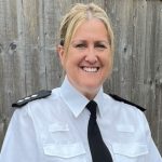 Law enforcement officer in white uniform shirt with black epaulets and black tie stands before wooden fence