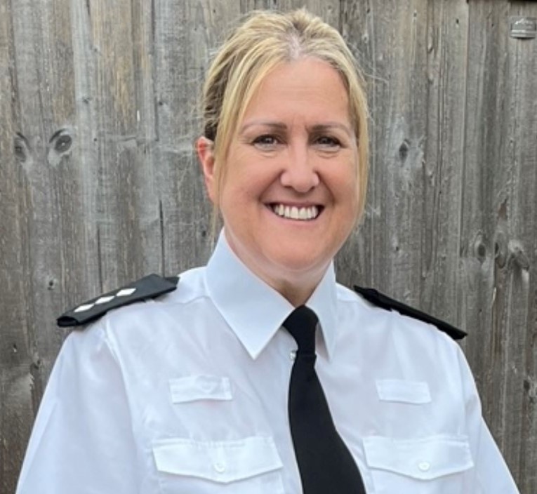 Law enforcement officer in white uniform shirt with black epaulets and black tie stands before wooden fence