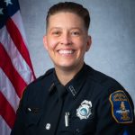 Police officer in dark uniform with badge, nameplate, and sergeant chevrons on sleeve, standing before U.S. flag and gray backdrop in formal portrait.