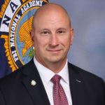 Person in dark suit with white shirt and patterned red tie stands before FBI National Academy flag featuring eagle emblem and blue-gray studio backdrop