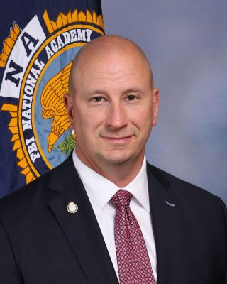 Person in dark suit with white shirt and patterned red tie stands before FBI National Academy flag featuring eagle emblem and blue-gray studio backdrop