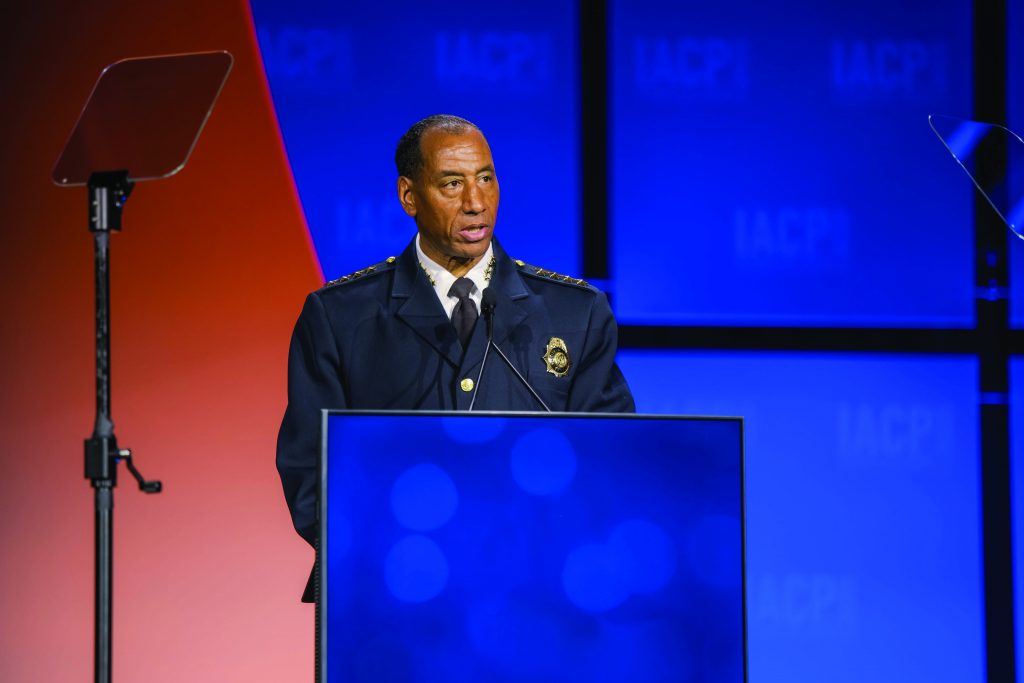 Uniformed law enforcement official stands at a podium with microphones on a stage featuring blue and orange panels and teleprompters positioned on both sides