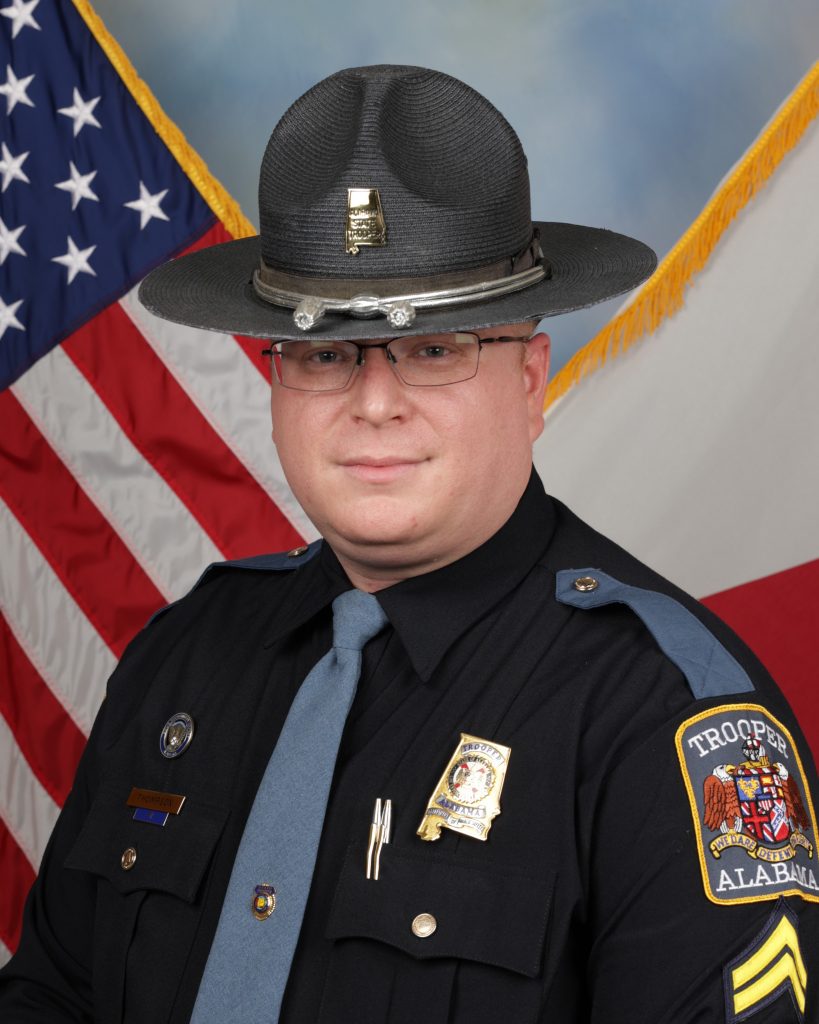 Formal studio portrait of a uniformed state trooper wearing a campaign hat, tie, badge, nameplate, and Alabama shoulder patch, posed before U.S. and state flags
