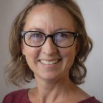 Individual with shoulder-length wavy hair, gold hoop earrings, and maroon top stands against a plain background