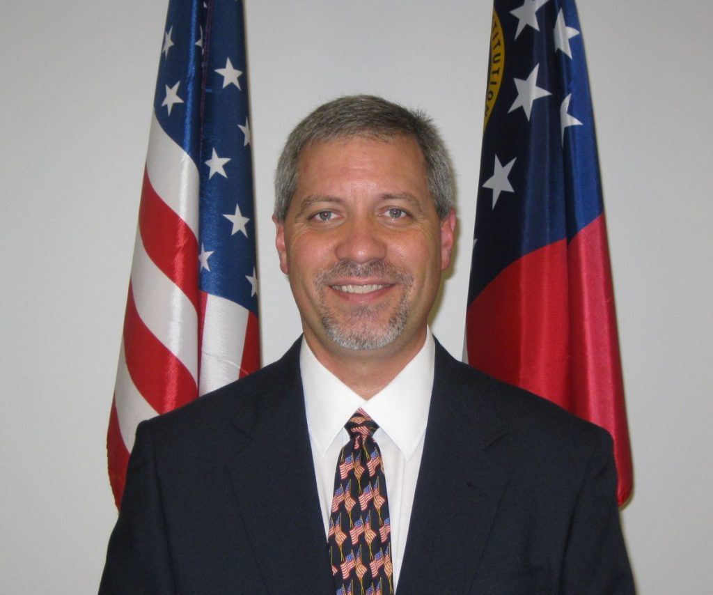 Individual in dark suit, white shirt, and American flag tie stands before U.S. flag and state flag; formal setting with both flags fully visible behind
