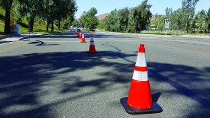 Orange traffic cones form a straight lane on a broad roadway, guiding vehicles past trees, sidewalks, utility poles, and signs in a well‑lit outdoor setting
