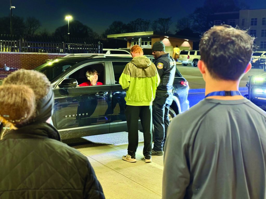 Police officer and person in green jacket stand beside black SUV at night, engaging with driver; three bystanders observe nearby.