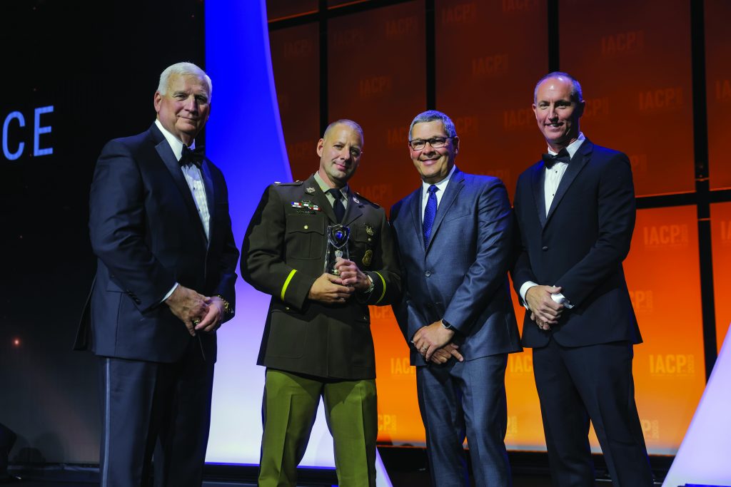 Four individuals in formal attire stand on stage with orange and purple backdrop; one in brown military-style uniform holds a glass award