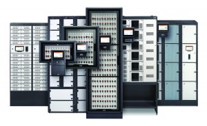 The image shows a variety of electronic control panels and cabinets arranged in a row. There are seven main units, each with multiple smaller components and displays. The central unit is the largest, featuring numerous small switches or indicators arranged in a grid pattern. Surrounding this central unit are six other panels of varying sizes, each with similar but distinct configurations of switches, buttons, and digital displays. The overall color scheme is predominantly gray and black with some orange accents on the switches or indicators.