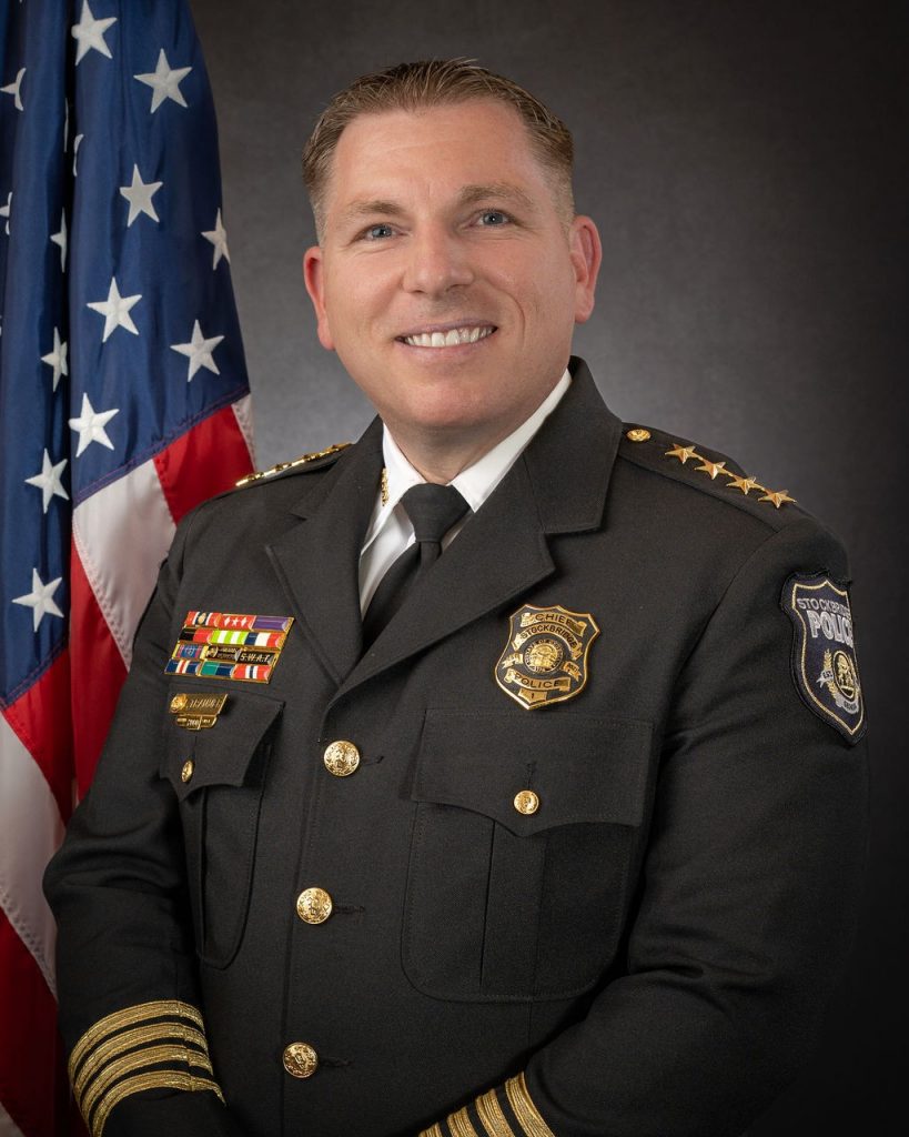 Uniformed law enforcement officer in dark jacket with gold buttons, badges, and shoulder insignia stands before American flag on plain background.