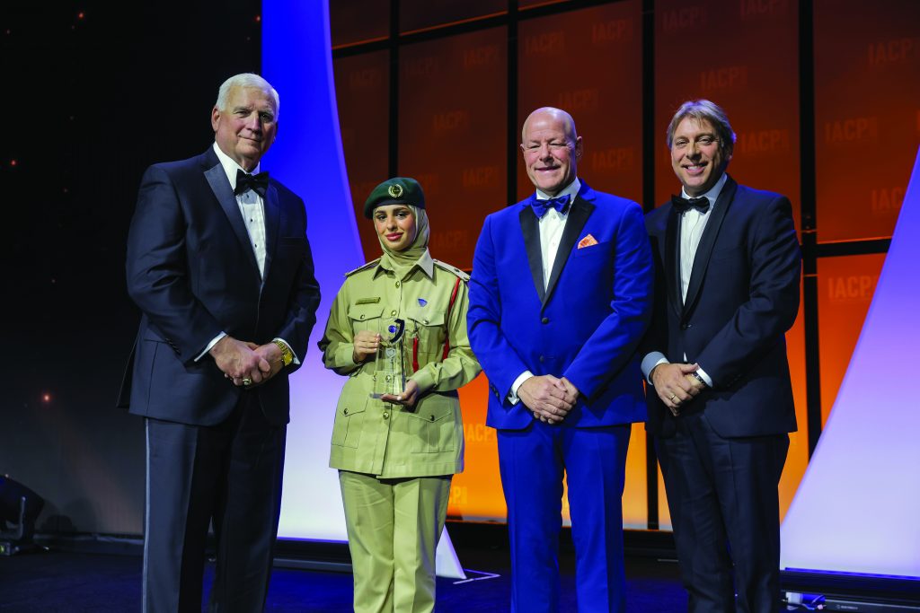 Four individuals stand on stage with orange and purple backdrop; one in tan military-style uniform and green beret holds a glass award, others in formal suits