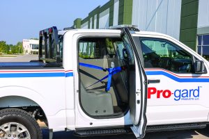White Pro-gard pickup truck parked outside a modern building with rear door open, showing blue restraint system and partition installed inside the cab