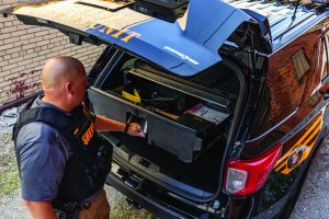 Sheriff’s deputy opens a rear storage drawer in a patrol SUV, revealing organized gear, equipment trays, and documents under the raised hatch beside a brick wall