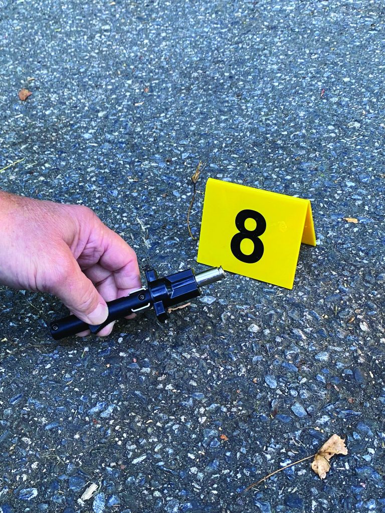 Hand holds a black evidence tool near a yellow marker labeled 8 on rough asphalt scattered with small debris during outdoor crime‑scene documentation