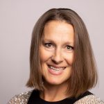 Individual with shoulder-length brown hair wears black top with silver shoulder embellishments, standing against a plain light gray background