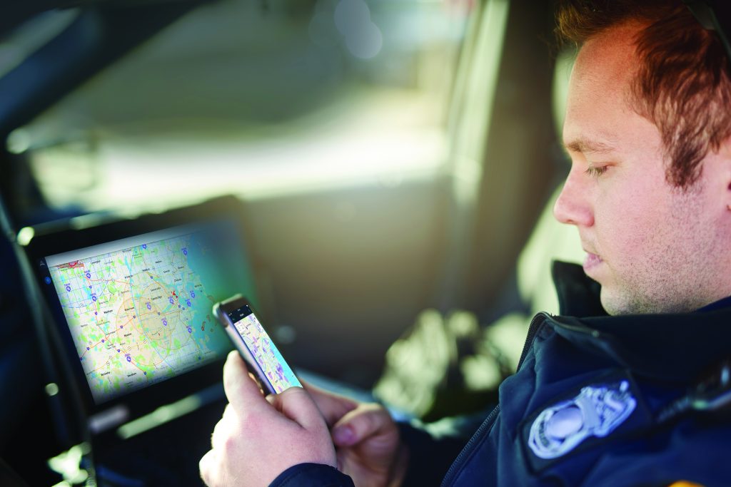 A police officer is seated inside a patrol car. The officer is wearing a dark uniform with an embroidered badge on the left side of the chest. The officer holds a smartphone in the left hand, displaying a map application with various colored routes and markers. In front of the officer, mounted on the dashboard, is a larger screen showing a detailed map with similar routes and markers. Sunlight filters into the vehicle from outside, illuminating parts of the interior. This image shows a law enforcement officer using technology to navigate or monitor locations inside a patrol car.
