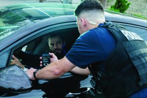 Officer in tactical vest marked POLICE shows a phone to a driver through an open car window, with radio earpiece, duty gear and dark sedan exterior