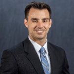 Individual in dark suit jacket, white dress shirt, and blue striped tie stands before plain dark background.