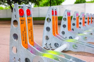 Rows of galvanized steel vehicle barriers with circular cutouts, crossbars, red reflectors, and orange Traffic Devices Inc. panels are aligned on pavement, with trees, grass, a concrete wall, and shallow depth of field visible in the background