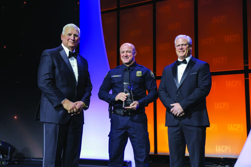 Three individuals stand on stage with orange and purple backdrop; one in dark police uniform holds a glass award, flanked by two in black tuxedos