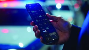 Hand holding a smartphone displaying an investigative app interface, with blurred police vehicle lights in red and blue illuminating the background