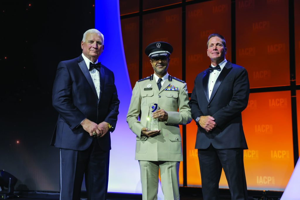Three individuals stand on stage with orange and purple backdrop; one in tan formal uniform with medals and peaked cap holds a glass award, others in tuxedos