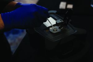 Gloved hand uses tool to handle sample on forensic device, highlighting analytical equipment and precise evidence processing procedure.