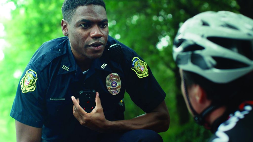 Uniformed officer with badges and body camera kneels beside helmeted cyclist on path; background features dense green foliage and natural setting