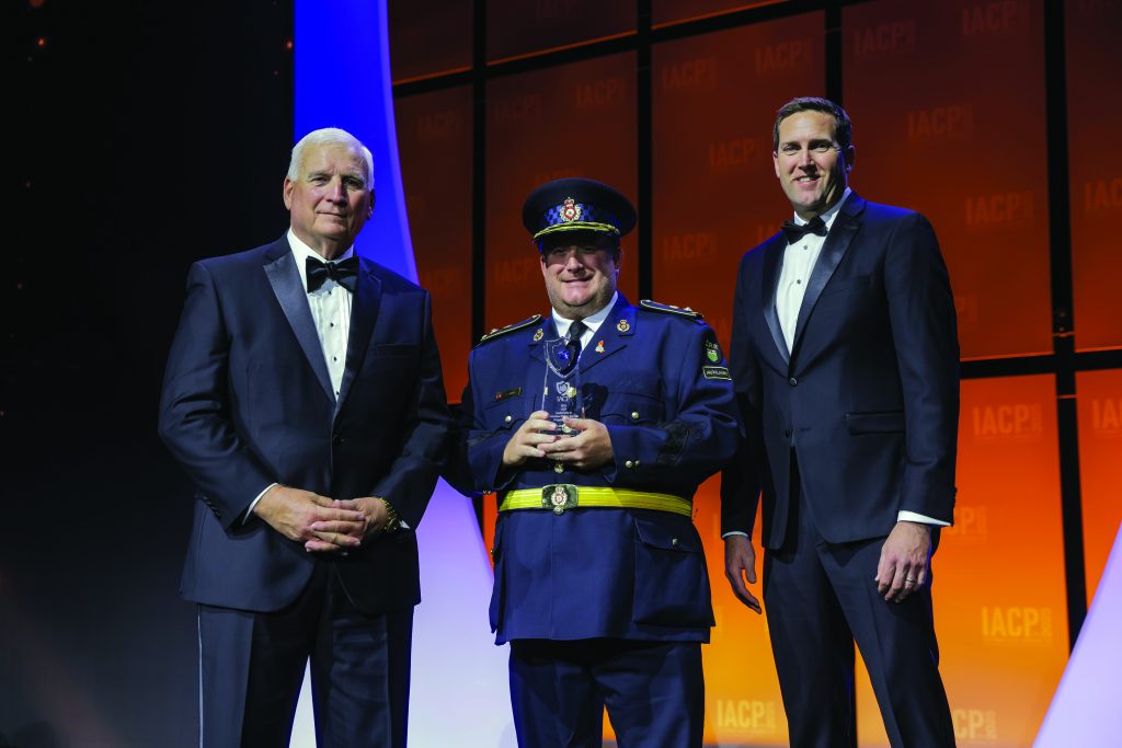 Three individuals stand on stage with orange and purple backdrop; one in dark blue formal uniform with peaked cap and yellow belt holds a glass award