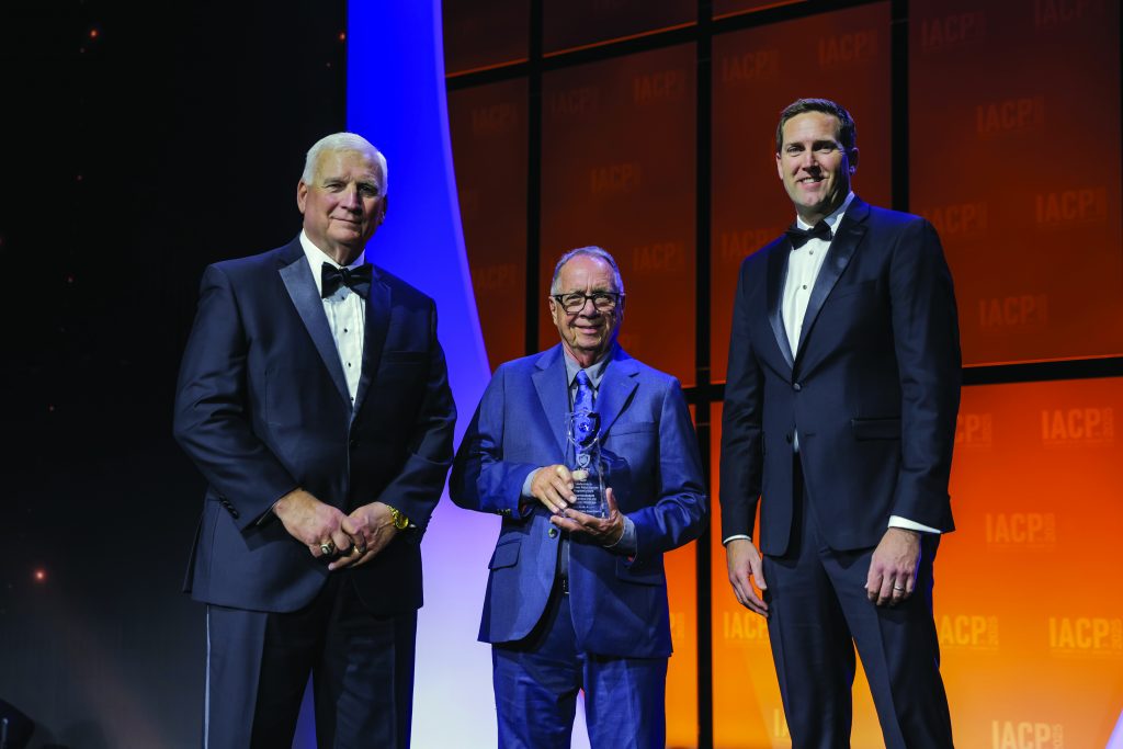 Three individuals stand on stage with orange and purple backdrop; center person in blue suit holds a glass award, flanked by two in black tuxedos