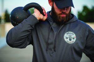 A person is holding a black kettlebell on the right shoulder with the right hand. The person is wearing a gray hoodie with a circular logo on the left chest area. The logo has "WELLFIT" written at the top, two crossed barbells in the center, and "EST 2018" at the bottom. The background appears to be an outdoor setting with blurred greenery and structures.