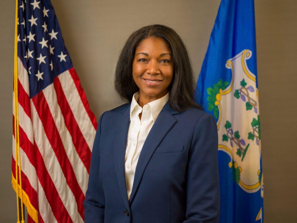 Person in navy blazer and white collared shirt standing between U.S. flag and blue state flag with emblem, against a neutral indoor background