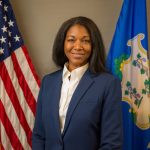 Person in navy blazer and white collared shirt standing between U.S. flag and blue state flag with emblem, against a neutral indoor background