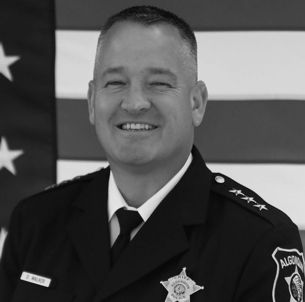A black and white photograph of a police officer standing in front of an American flag. The officer is wearing a formal police uniform, including a dark jacket with several buttons, epaulets with stripes indicating rank, and a badge on the left chest. The badge reads "Sheriff" and has a star shape. On the right chest, there is a name tag. The officer's arms are crossed in front of the body at waist level. The shoulder patch on the left arm reads "Madison Police."